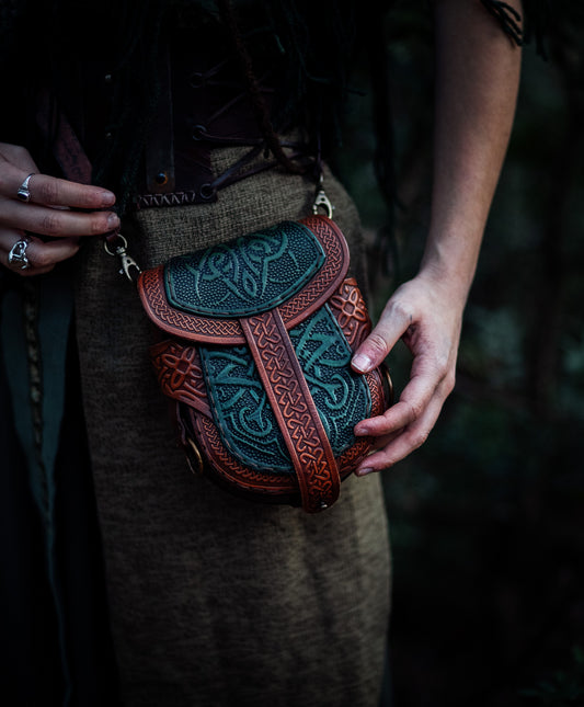 Close-up of a Celtic leather pouch held by both hands. The green embossed panels and brown leather frame create a rich fantasy look. The detailed craftsmanship makes it ideal for immersive LARP characters or medieval-themed daily wear. The pouch is secured with a leather strap and can be worn with or without a shoulder strap. A standout accessory for anyone seeking authenticity and elegance.