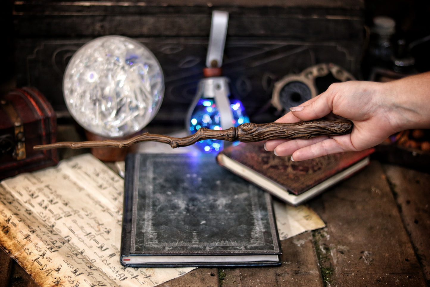 A hand holding a twisted branch-shaped magic wand with a blue light, placed on an open book in a dark room with a mirror and other magical items. Ancient Branch Magic Wand: Enchanting Elegance for LARP, Ren-Fair, Wizardry, Sorcery, Witchcraft, and Fairy Realms - Handcrafted Marvel