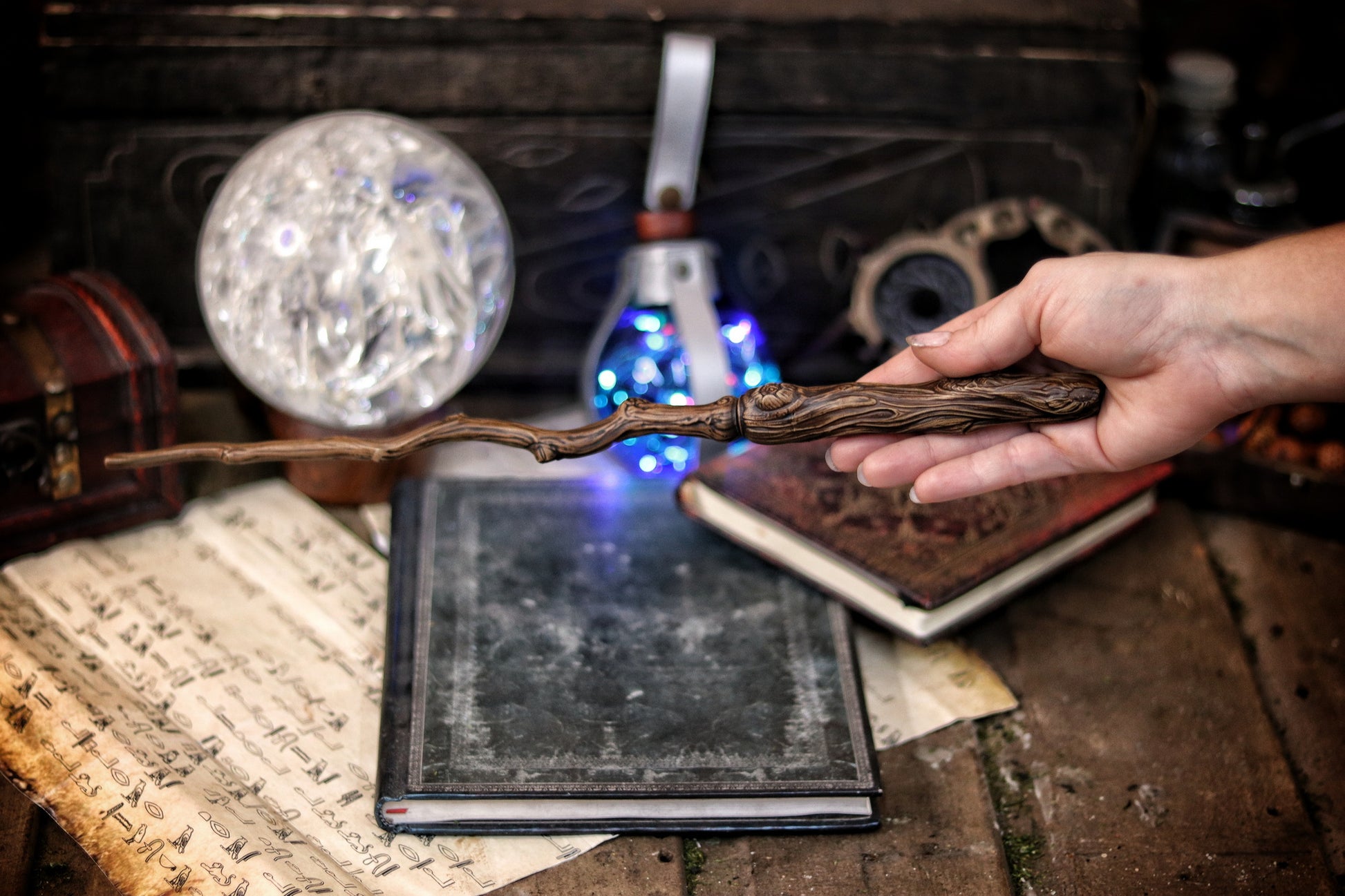 A hand holding a twisted branch-shaped magic wand with a blue light, placed on an open book in a dark room with a mirror and other magical items. Ancient Branch Magic Wand: Enchanting Elegance for LARP, Ren-Fair, Wizardry, Sorcery, Witchcraft, and Fairy Realms - Handcrafted Marvel