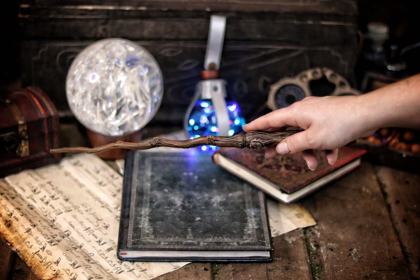 A hand holding a twisted branch-shaped magic wand with a blue light, placed on an open book in a dark room with a mirror and other magical items. Ancient Branch Magic Wand: Enchanting Elegance for LARP, Ren-Fair, Wizardry, Sorcery, Witchcraft, and Fairy Realms - Handcrafted Marvel