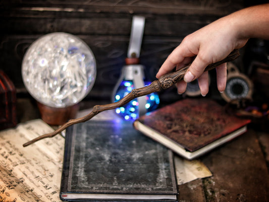 A hand holding a twisted branch-shaped magic wand with a blue light, placed on an open book in a dark room with a mirror and other magical items. Ancient Branch Magic Wand: Enchanting Elegance for LARP, Ren-Fair, Wizardry, Sorcery, Witchcraft, and Fairy Realms - Handcrafted Marvel