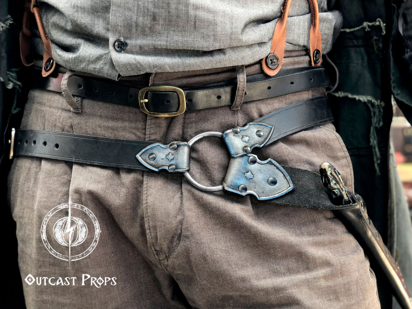 Person wearing a detailed belt with a large buckle, likely from a historical or fantasy costume, with 'Outcast Props' branding. Waist-level shot of a leather wand holster belt styled with a long coat, ideal for dark wizard costumes. Great for Death Eater cosplay, wizard LARP, or gothic fantasy costumes. Combines functionality and style for Harry Potter-themed events and wizard duels.