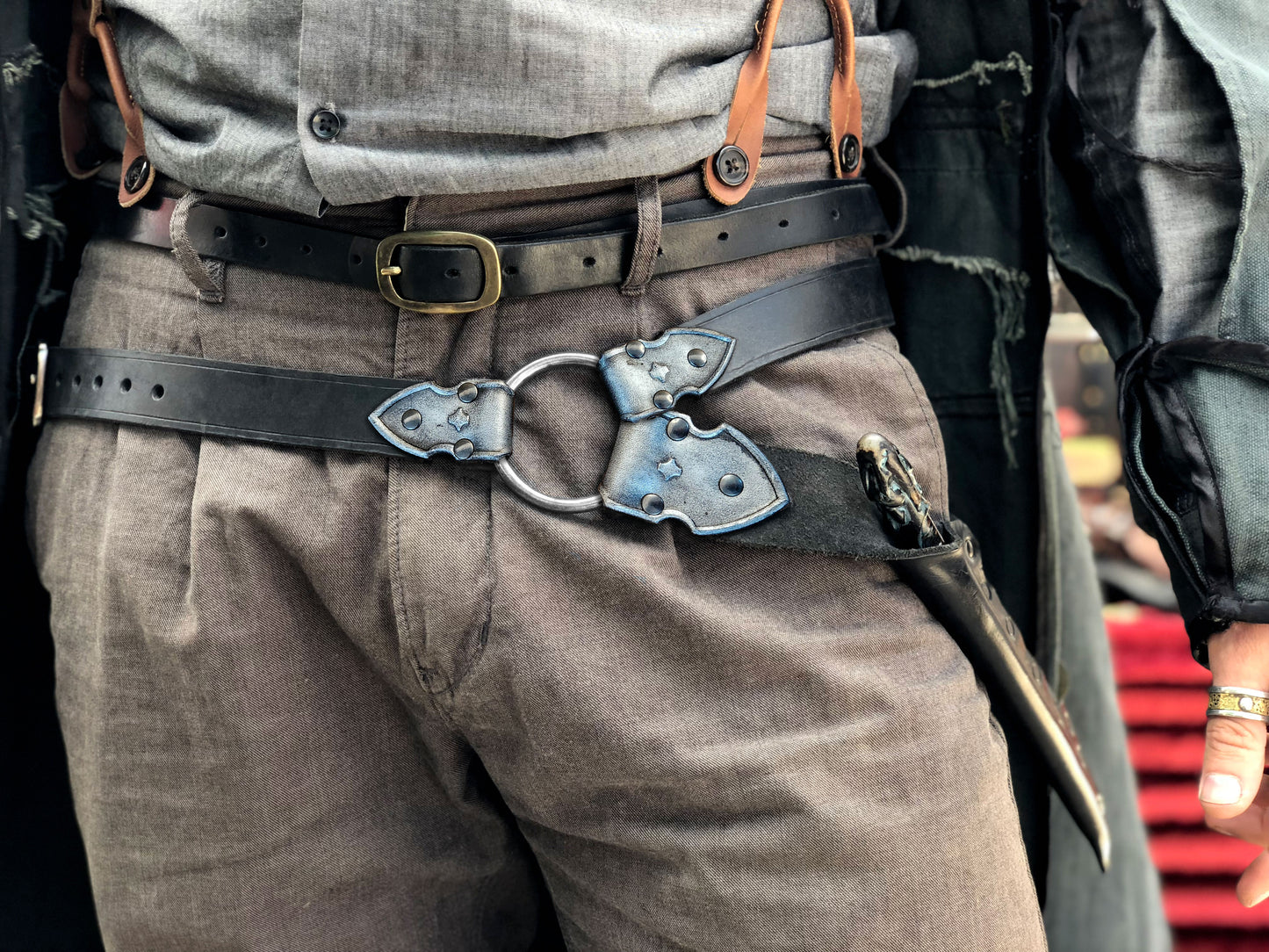 Waist-level shot of a leather wand holster belt styled with a long coat, ideal for dark wizard costumes. Great for Death Eater cosplay, wizard LARP, or gothic fantasy costumes. Combines functionality and style for Harry Potter-themed events and wizard duels. Person wearing a black leather harness on gray pants with a blurred background. Person wearing a detailed belt with a large buckle, gray pants, and a jacket.