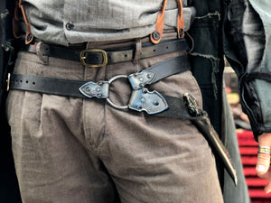 Waist-level shot of a leather wand holster belt styled with a long coat, ideal for dark wizard costumes. Great for Death Eater cosplay, wizard LARP, or gothic fantasy costumes. Combines functionality and style for Harry Potter-themed events and wizard duels. Person wearing a black leather harness on gray pants with a blurred background. Person wearing a detailed belt with a large buckle, gray pants, and a jacket.