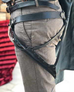 Waist-level shot of a leather wand holster belt styled with a long coat, ideal for dark wizard costumes. Great for Death Eater cosplay, wizard LARP, or gothic fantasy costumes. Combines functionality and style for Harry Potter-themed events and wizard duels. Person wearing a black leather harness on gray pants with a blurred background