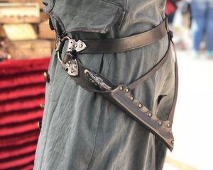 Waist-level shot of a leather wand holster belt styled with a long coat, ideal for dark wizard costumes. Great for Death Eater cosplay, wizard LARP, or gothic fantasy costumes. Combines functionality and style for Harry Potter-themed events and wizard duels. Person wearing a black leather harness on gray pants with a blurred background. Gray costume with a belt and sword on a blurred background