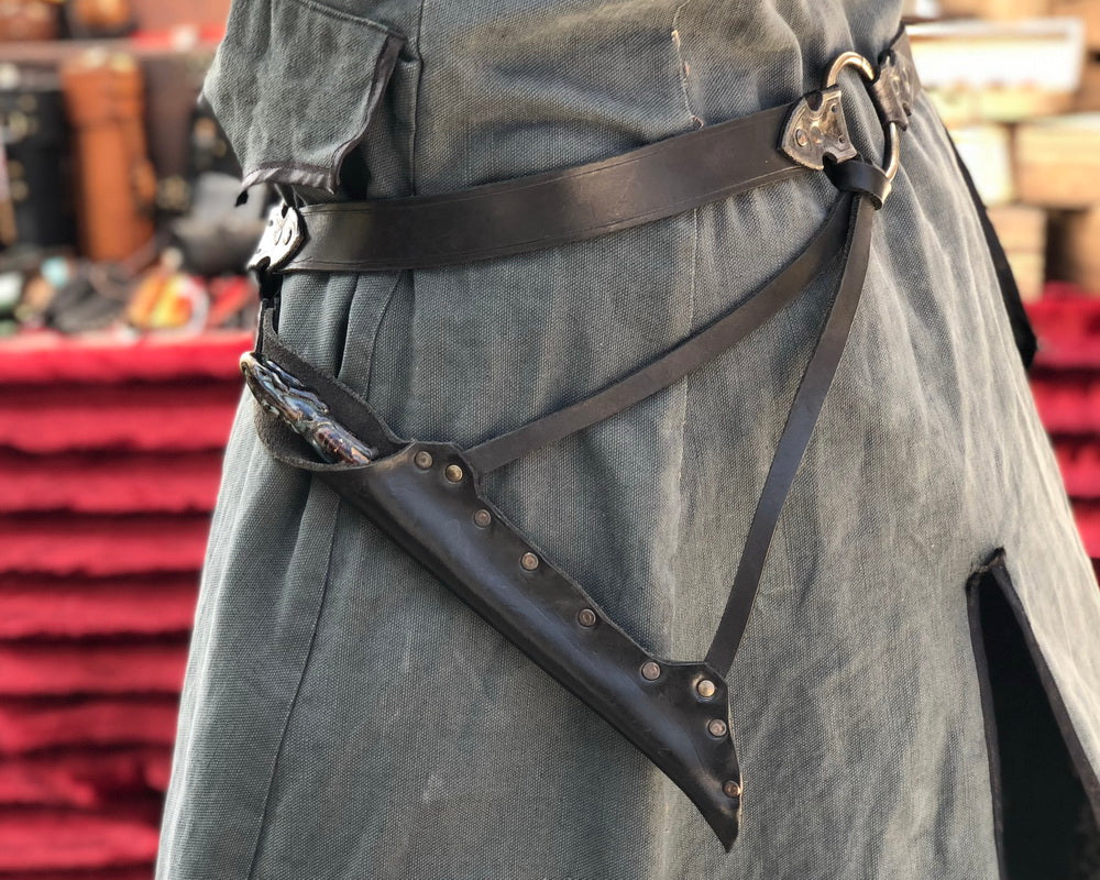 Waist-level shot of a leather wand holster belt styled with a long coat, ideal for dark wizard costumes. Great for Death Eater cosplay, wizard LARP, or gothic fantasy costumes. Combines functionality and style for Harry Potter-themed events and wizard duels. Person wearing a black leather harness on gray pants with a blurred background. Person wearing a dark leather harness with metal studs against a blurred background