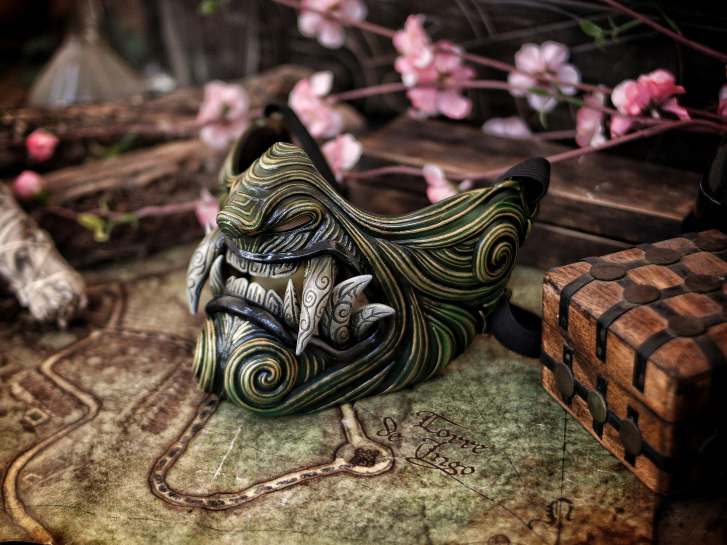 A hand-painted Oni Samurai Mempo mask with gold, green and black details, displayed on a surface with a map background and pink flowers in the peripheral. Samurai oni mask japanese half oni mask samurai wearable mask for cosplay larp menpo mask scorpion clan mask l5r samurai mask