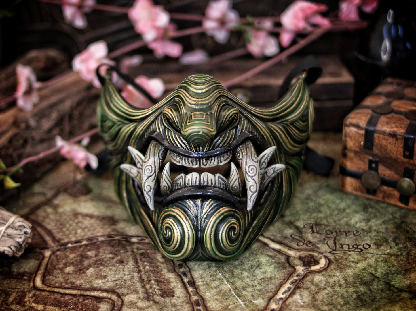 A Oni Samurai Mempo mask with green and black colors, featuring hand-painted details and a fierce design, displayed on a surface with a map and flowers in the background. Samurai oni mask japanese half oni mask samurai wearable mask for cosplay larp menpo mask scorpion clan mask l5r samurai mask