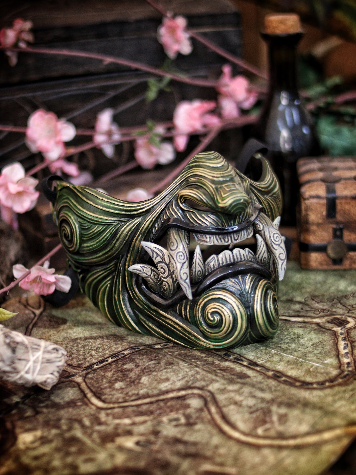 A Oni Samurai Mempo mask with green and black colors, featuring hand-painted details and a fierce design, displayed on a surface with a map and flowers in the background. Samurai oni mask japanese half oni mask samurai wearable mask for cosplay larp menpo mask scorpion clan mask l5r samurai mask