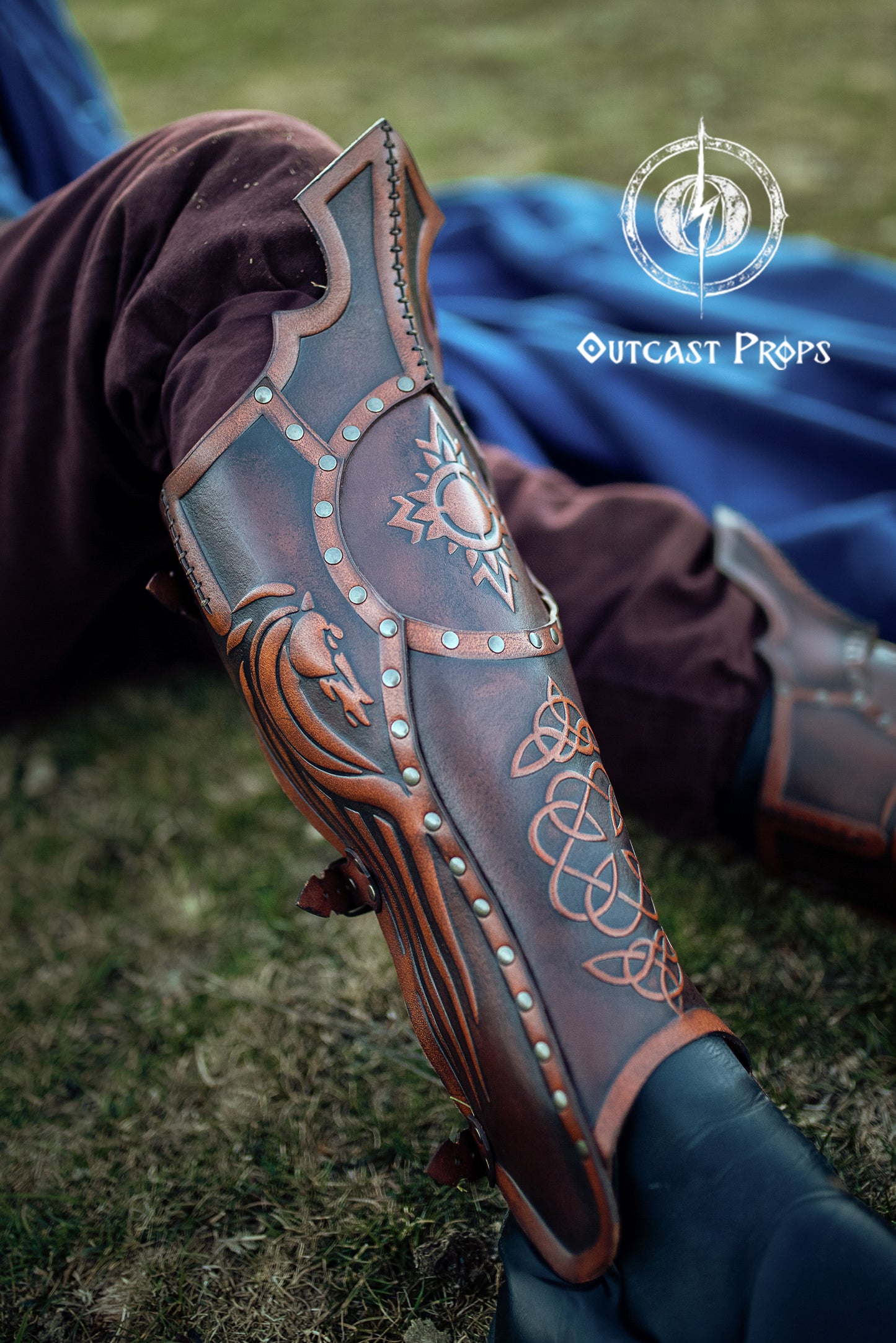 A LARP warrior lies on grass wearing dark reddish-brown leather greaves embossed with Celtic knots, a dragon, and a radiant sun. The leg armor is secured with straps and rivets, and shaped with a pointed top over the knee. The embossed motifs are orange-highlighted, contrasting with the aged leather finish. In the background, a deep blue cloak and burgundy pants create a vivid fantasy costume. Ideal for Viking, medieval, or Celtic cosplay and LARP. Celtic Leather Greaves, medieval armor for LARP or cosplay,