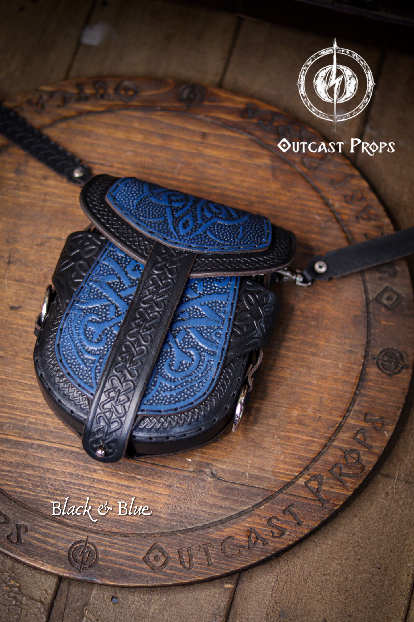 Black and blue celtic leather bag with intricate designs on a wooden surface, branded 'Outcast Props'. Celtic bag Leather Black and Blue