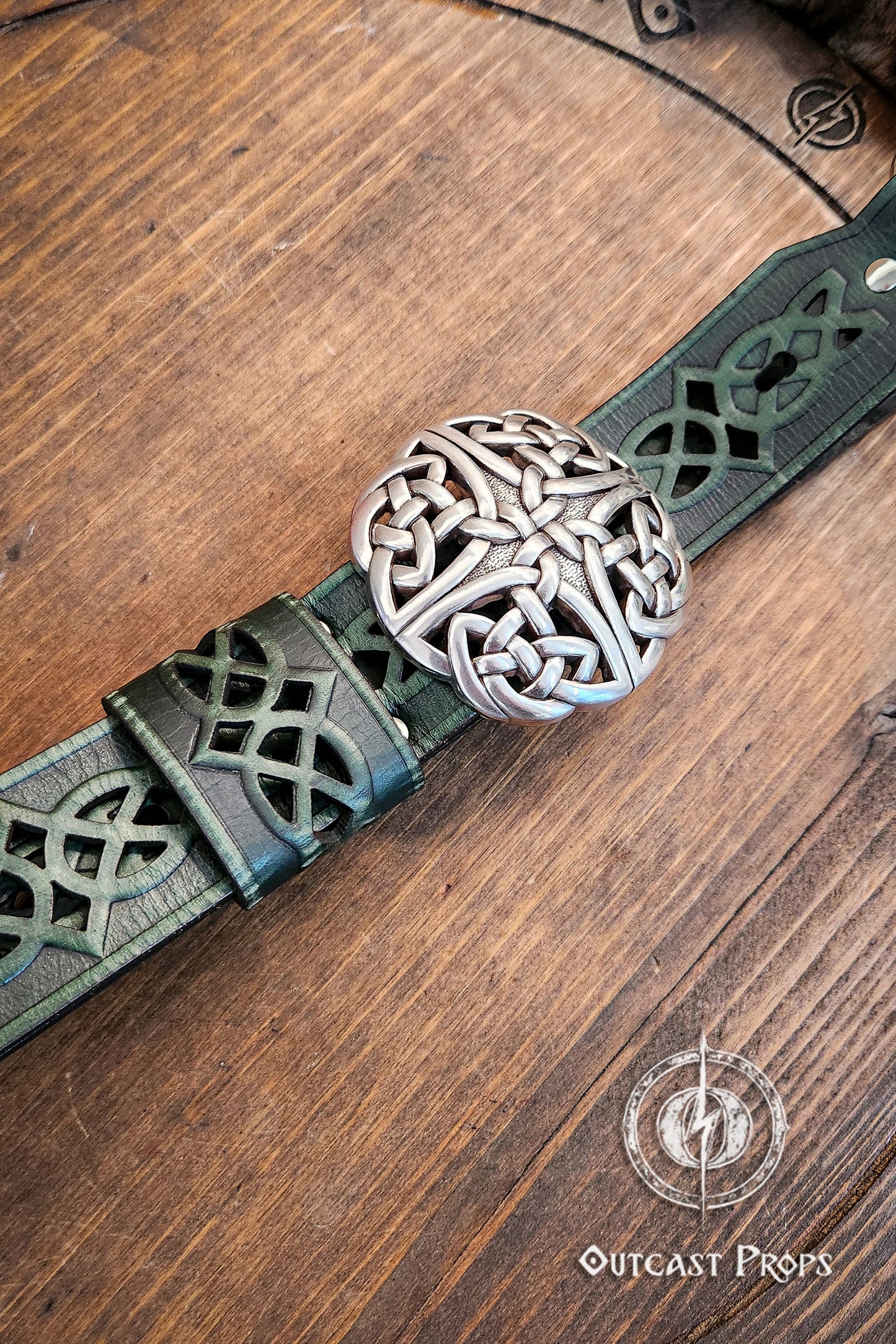 Green leather belt with silver Celtic knot buckle on a wooden surface, branded 'Outcast Props'. Celtic knot leather belt silver buckle larp belt for ren fair fantasy costume celtic belt unisex leather belt for renaissance outfit