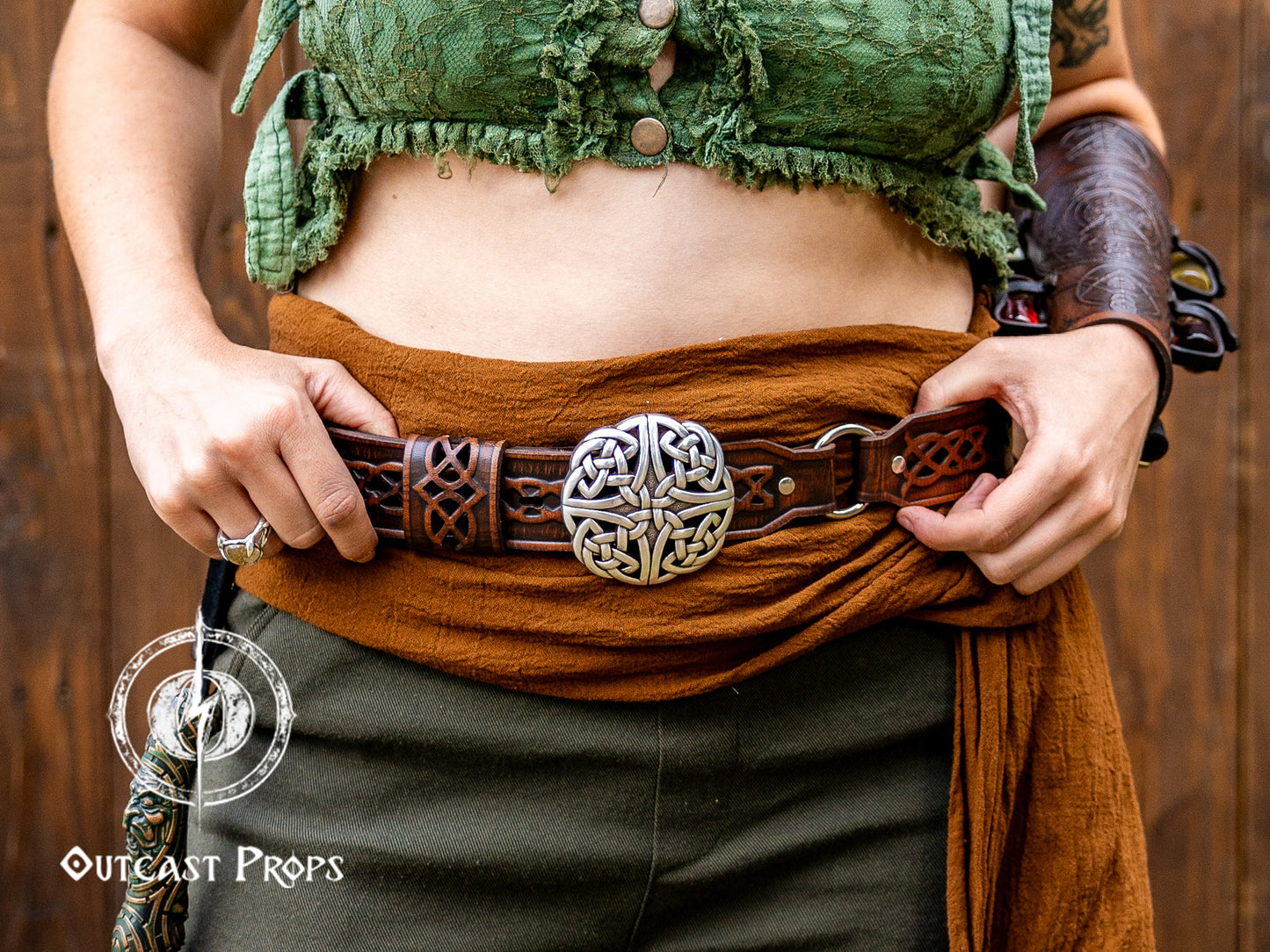 Person wearing a green top and brown belt with a decorative buckle against a wooden background, with 'Outcast Props' logo. Celtic knot leather belt silver buckle larp belt for ren fair fantasy costume celtic belt unisex leather belt for renaissance outfit