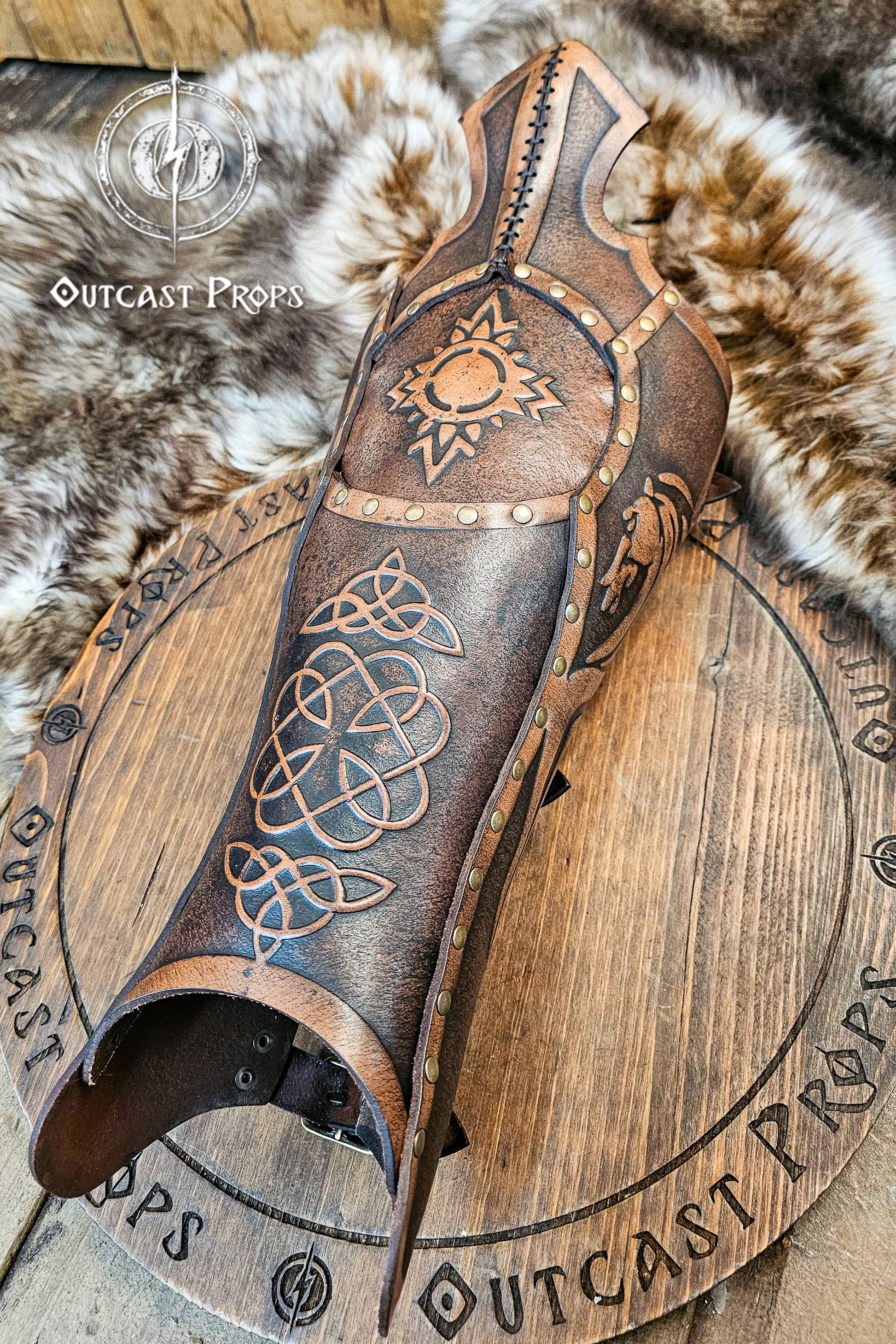 Single handcrafted Celtic leather greave resting on fur, embossed with knotwork and solar symbols recalling the heritage of Rohirrim riders and Viking leg armor. Made from vegetable-tanned leather aged by hand, with brass rivets and sculpted edges. Perfect for fantasy LARP, horsemen, and medieval reenactment costumes seeking authentic, eco-friendly craftsmanship and the distinctive aged finish of Outcast Props.