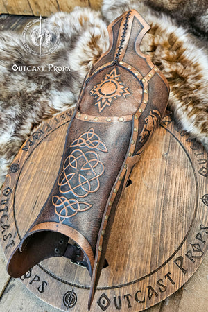 Single handcrafted Celtic leather greave resting on fur, embossed with knotwork and solar symbols recalling the heritage of Rohirrim riders and Viking leg armor. Made from vegetable-tanned leather aged by hand, with brass rivets and sculpted edges. Perfect for fantasy LARP, horsemen, and medieval reenactment costumes seeking authentic, eco-friendly craftsmanship and the distinctive aged finish of Outcast Props.