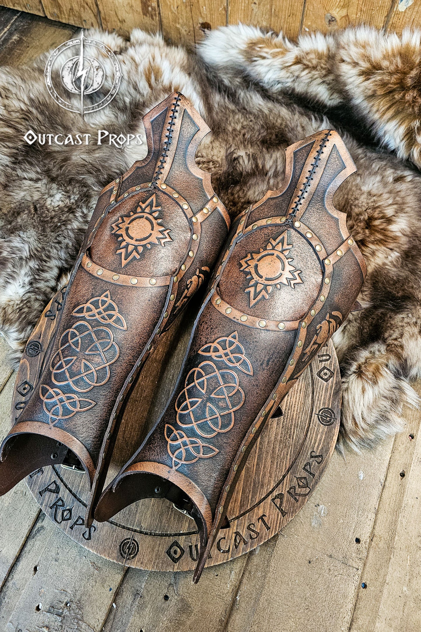 Pair of Celtic leather greaves resting on fur background, hand-embossed with triquetra and sun patterns reminiscent of Rohirrim and Viking heritage. Crafted in aged brown vegetable-tanned leather, riveted and dyed by hand for LARP, cosplay, or medieval horsemen costumes. A perfect combination of protection and artistry for fantasy warriors seeking authentic craftsmanship and cinematic detailing by Outcast Props.