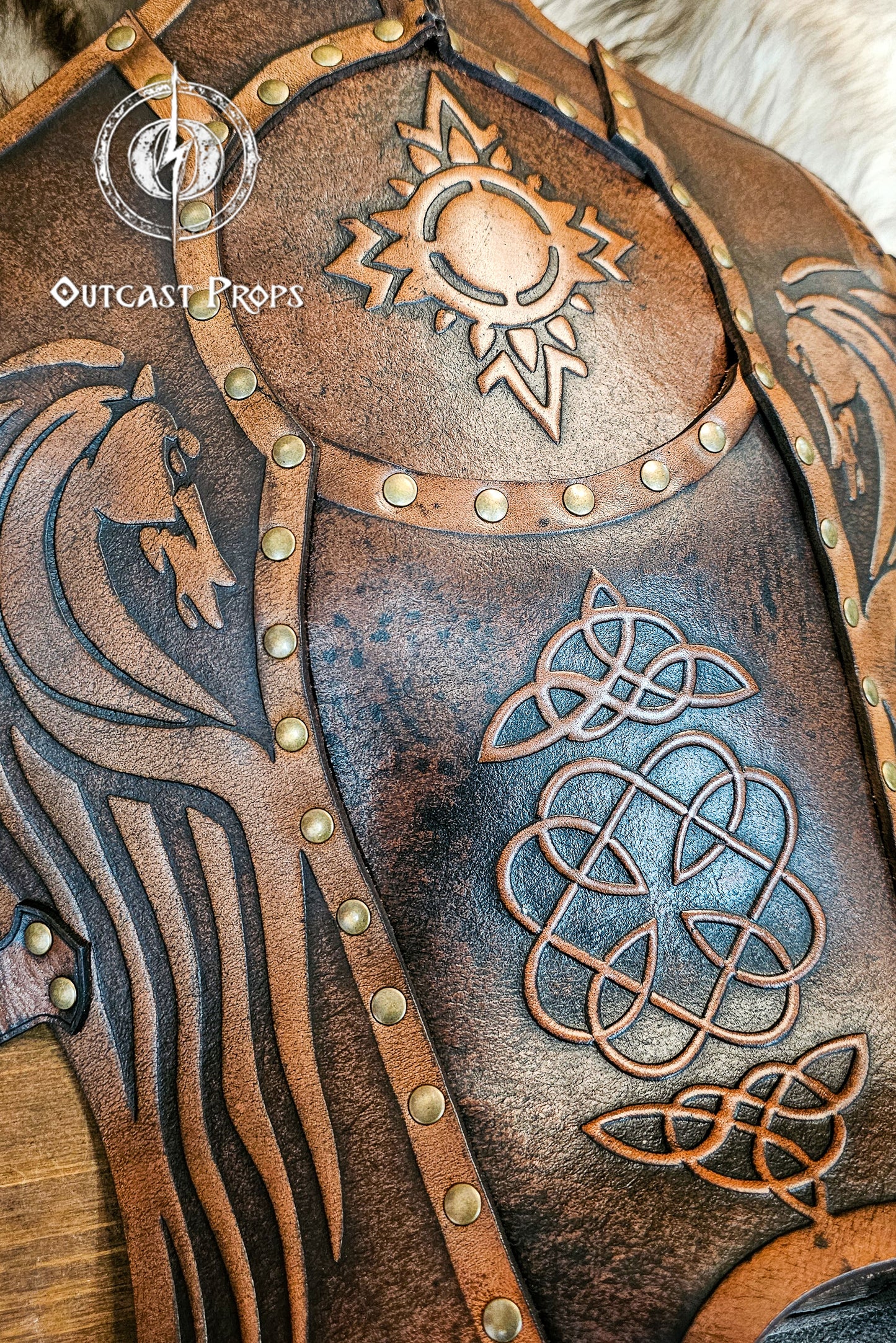 Detailed close-up of handcrafted Celtic leather greaves featuring embossed sun and knotwork designs with twin horse motifs reminiscent of Rohirrim armor. The brown full-grain leather surface, aged with natural dyes and brass rivets, gives a warm, battle-worn look. Ideal for LARP, Viking, and medieval horsemen cosplay, combining protection, heritage, and cinematic quality through Outcast Props’ unique aging techniques.