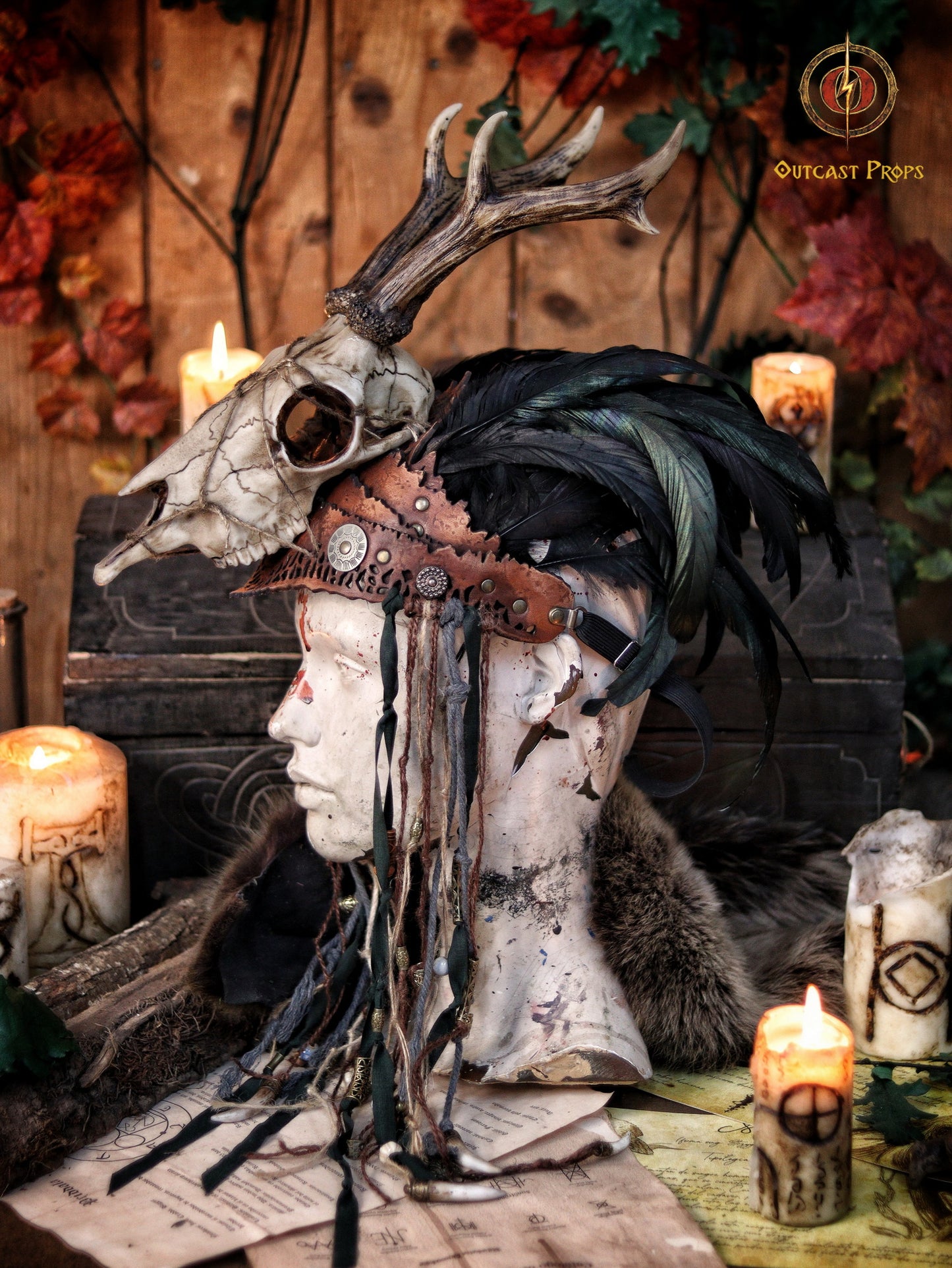 A Celtic adorned tiara with a resin skull, deer horns, feathers, bone pieces, beads, and fangs displayed on a table with a wooden background and candles. Beltane Headpiece tribal deer horns skull and feathers tiara leather and resin