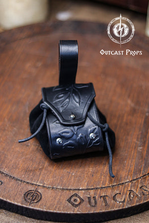 A black leather coin pouch with embossed botanical motifs stands on a circular wooden board engraved with the Outcast Props logo. The pouch combines a structured vegetable tanned leather flap with a soft suede body and silver rivets. Dark and elegant, it works perfectly for witches, shadowy elves, nobles or herbalist characters at a renfaire or LARP event. This fantasy dice pouch attaches to the belt and holds coins or small items securely.