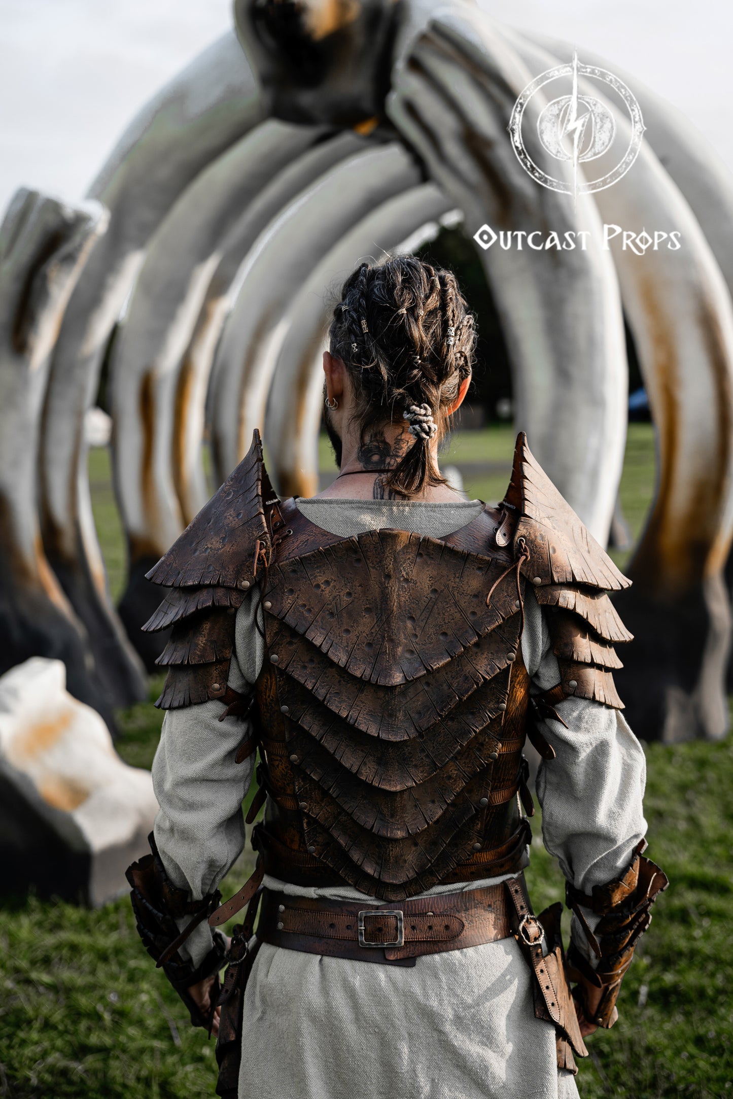 Back view of a warrior wearing the Dragon Scales Armor Set, standing before a giant bone-like sculpture at a LARP event. The armor is handmade from premium vegetable-tanned leather, textured to mimic ancient dragon hide. Shoulder guards and layered plates give a fierce, battle-ready look, ideal for cosplay, fantasy photoshoots, and immersive roleplay at Ren Faires or Viking festivals.
