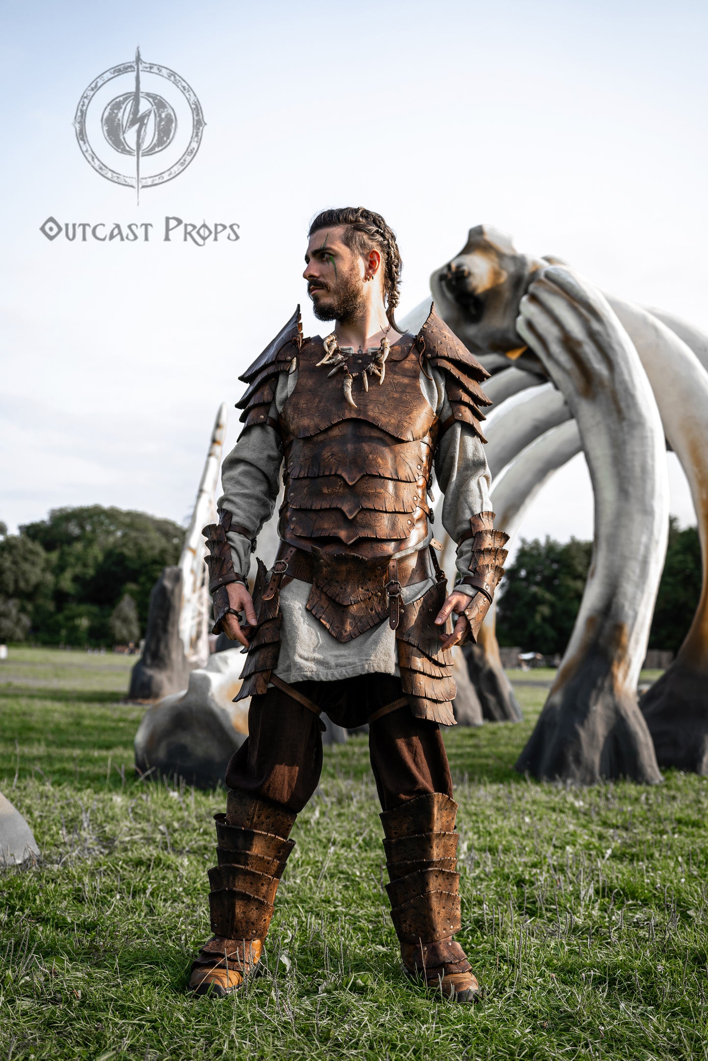 Full body photo of a LARP player dressed in the Dragon Scales Armor Set, in front of monumental bone structures. The complete leather outfit includes breastplate, pauldrons, arm guards, tassets, belt and greaves, all textured and dyed to resemble dragon hide. This fantasy armor is perfect for epic cosplay, Viking events, or fantasy reenactments. Made from eco-friendly leather and finished with Outcast Props’ signature techniques.
