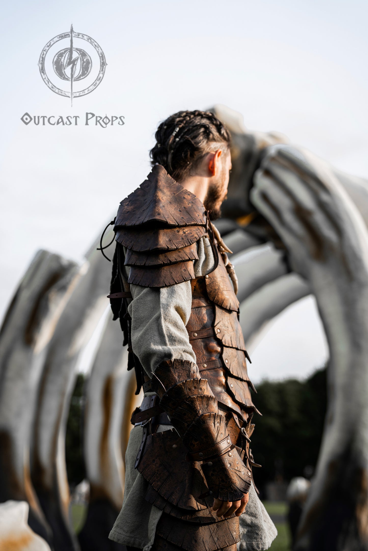 Close-up of the Dragon Scales armor set by Outcast Props, made from layered brown leather scales. Worn by a LARP character standing under giant bones, this fantasy armor includes a shoulder piece and bracer. Ideal for cosplay, fantasy events and renaissance fairs. Crafted with vegetable-tanned leather and a textured aged finish. Perfect for elves, warriors or druids.