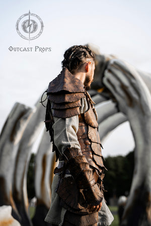 Close-up of the Dragon Scales armor set by Outcast Props, made from layered brown leather scales. Worn by a LARP character standing under giant bones, this fantasy armor includes a shoulder piece and bracer. Ideal for cosplay, fantasy events and renaissance fairs. Crafted with vegetable-tanned leather and a textured aged finish. Perfect for elves, warriors or druids.