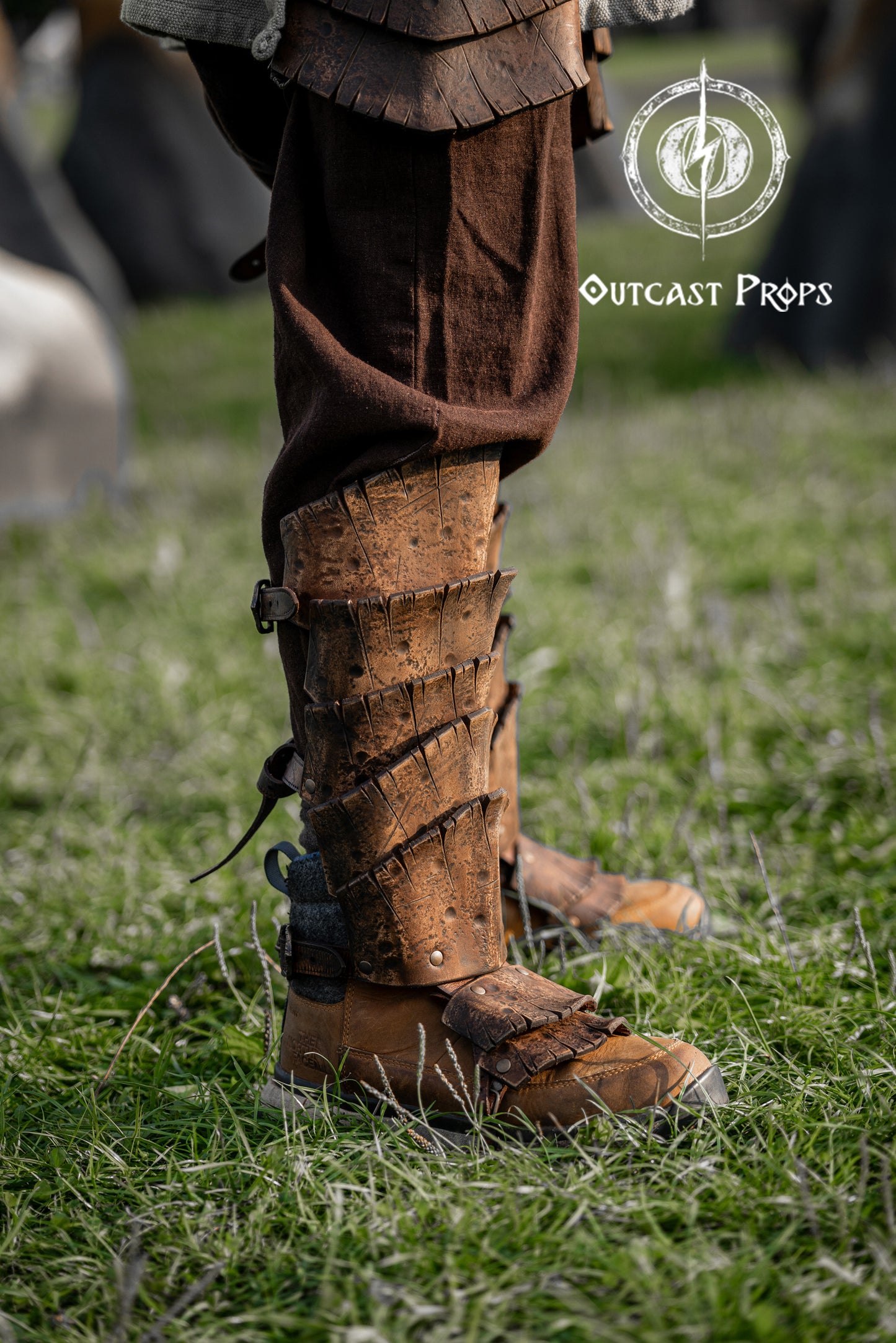 Close-up of handmade leather greaves inspired by dragon scales, worn over brown linen trousers and rustic boots on a grassy field. The greaves feature layered segments with deep textures and a rugged finish, ideal for fantasy costumes, LARP armor, or medieval warrior cosplay. Perfect for elves, rangers, or wild warriors attending Renaissance fairs. Detailed work showcases the artisanal leathercraft of Outcast Props.
