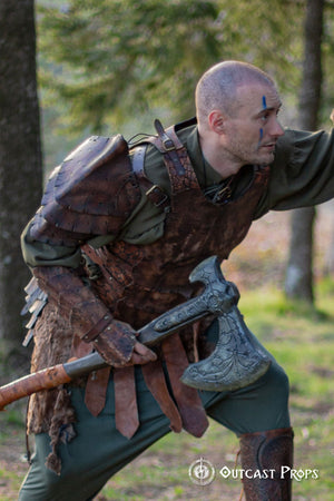 Portrait of a warrior with blue tribal face paint and layered dragon scale armor, including the matching leather bracer. Set in a mountain landscape, this shot highlights the bracer's texture and handcrafted details. Ideal for LARP costumes, medieval fantasy cosplay or orc armor sets. Each bracer is made with durable leather and detailed using traditional artisan techniques.