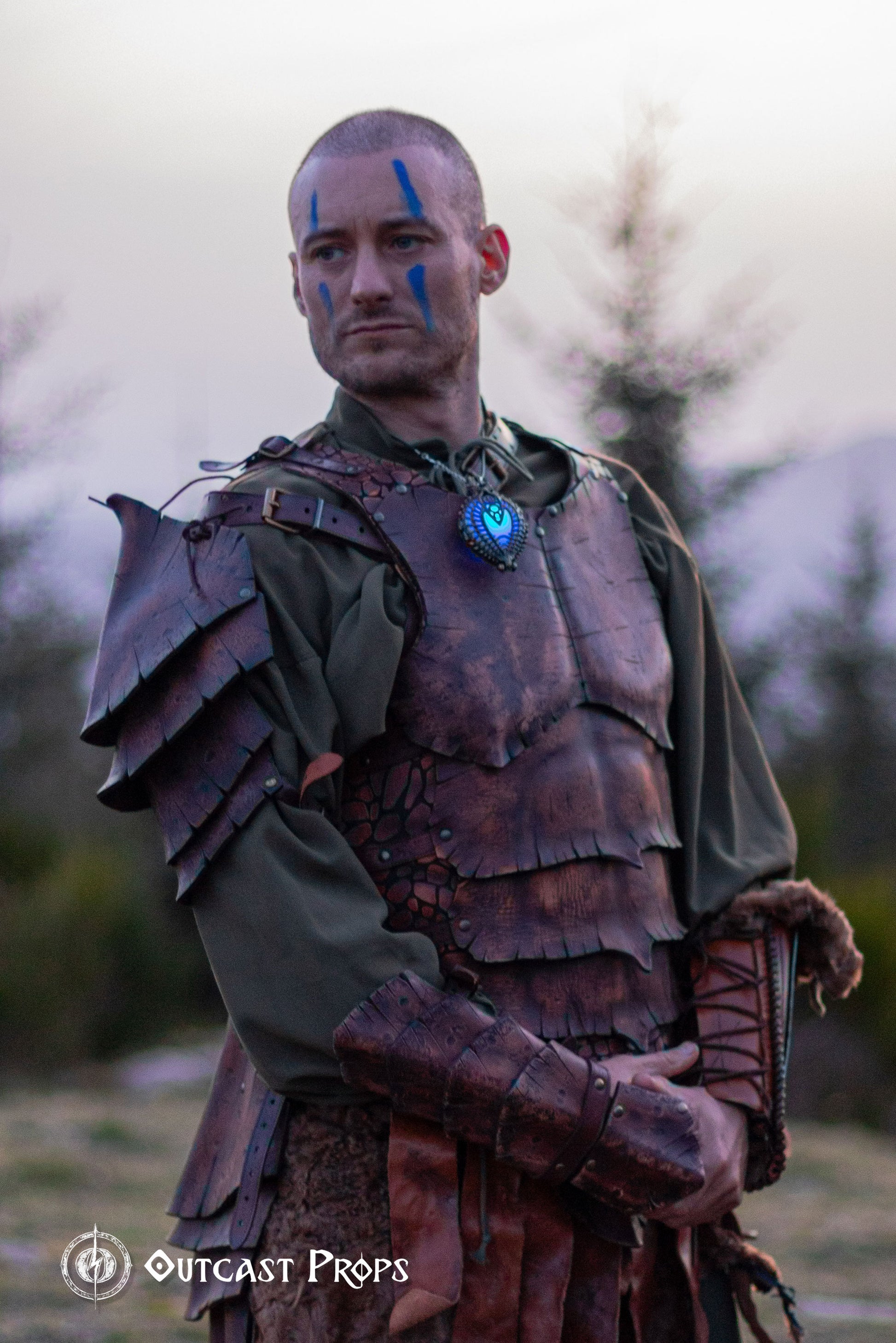 Portrait of a warrior with blue tribal face paint and layered dragon scale armor, including the matching leather bracer. Set in a mountain landscape, this shot highlights the bracer's texture and handcrafted details. Ideal for LARP costumes, medieval fantasy cosplay or orc armor sets. Each bracer is made with durable leather and detailed using traditional artisan techniques.