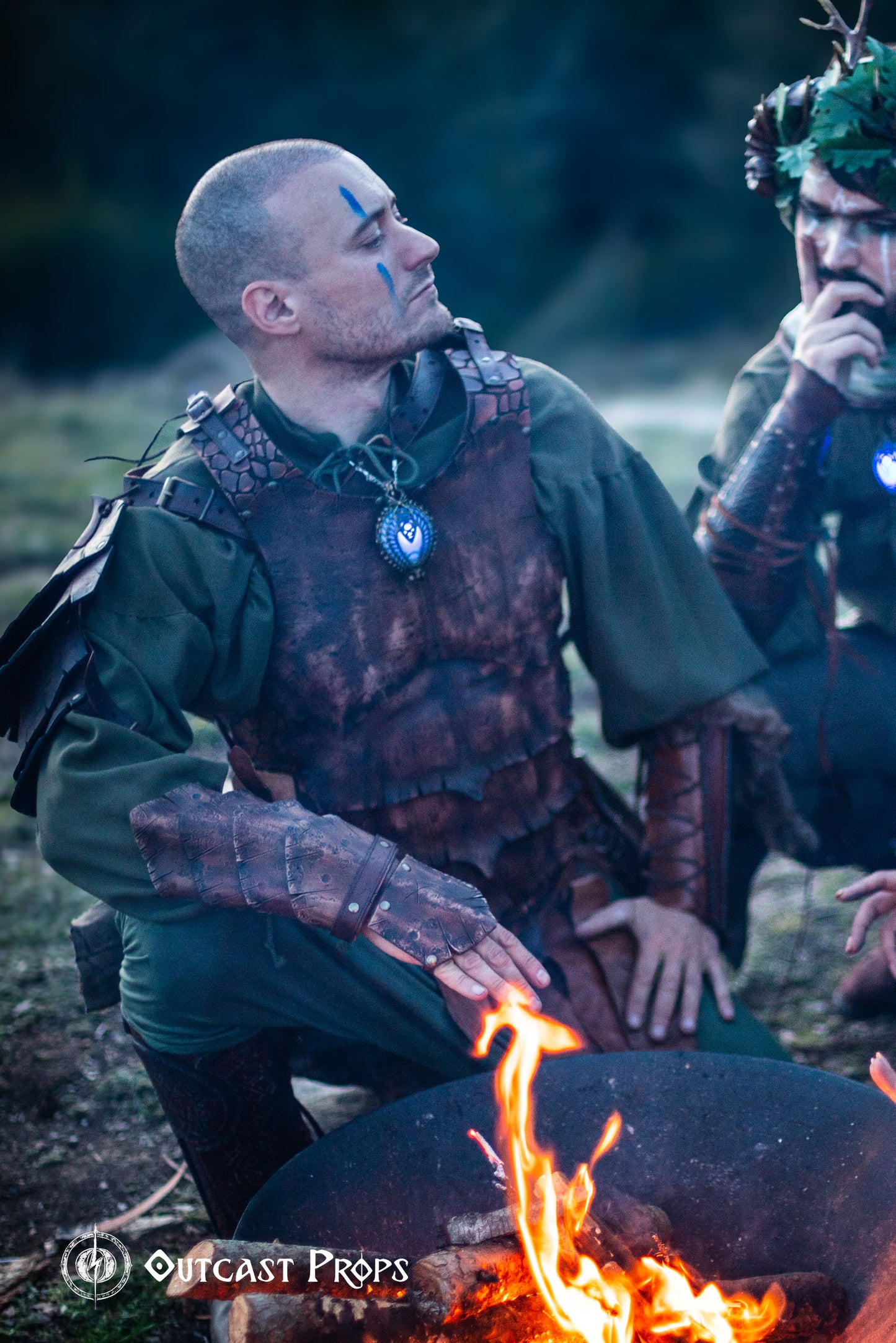 LARP warrior kneels by a fire, wearing a brown dragon scale leather bracer and matching armor with blue face paint. The bracer evokes strength and survival, perfect for orc cosplay, barbarian costumes or medieval fair outfits. Handcrafted with vegetable-tanned leather and hand-painted aging effects, this fantasy accessory brings gritty realism to your roleplay gear.