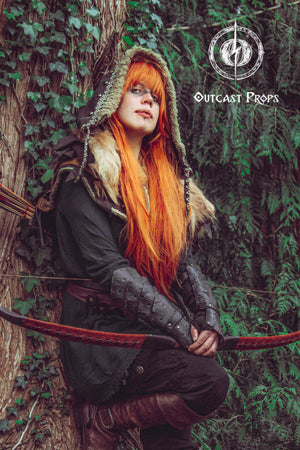 Female archer character with long orange hair and fur-trimmed hood leans against a tree, wearing black dragon scale bracers. The bracers add fierce elegance to LARP costumes, fantasy elf cosplay or rogue outfits. Made from genuine leather and aged by hand, these vambraces offer both comfort and bold style for medieval fairs or immersive fantasy adventures.