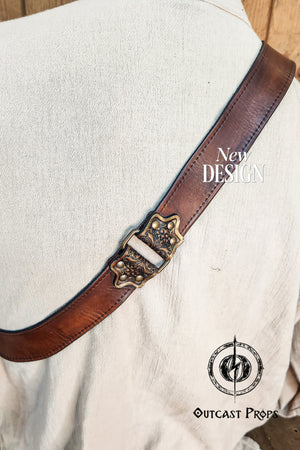 Edward Kenway pirate cosplay. Close-up of a handmade leather baldric strap resting diagonally across the back of a cream medieval tunic. The strap features premium brown vegetable-tanned leather and an ornate brass buckle with floral motifs. Ideal for pirate cosplay, steampunk outfits, or LARP characters. The detailed stitching and leather aging add to its realism and craftsmanship. Perfect accessory for historical reenactments or fantasy events.