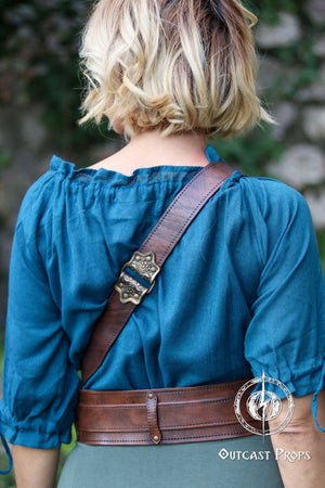 Edward Kenway pirate cosplay. A woman with short blond hair is shown from the back wearing a brown leather pirate baldric across a teal blouse. The leather strap curves diagonally over her shoulder and meets a wide waist belt, both decorated with stitching and an ornate brass buckle. The scene evokes pirate cosplay, renfaire outfits and noble adventurer aesthetics. The natural outdoor background adds realism, highlighting the aged texture of the leather baldric. baldric pirate belt flintlock