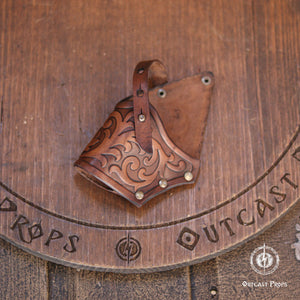 Brown leather holster featuring intricate embossed scrollwork patterns, a vertical strap with metal rivets, and a triangular shape. The leather has a polished finish with darker shading in the recessed areas of the decorative carvings.