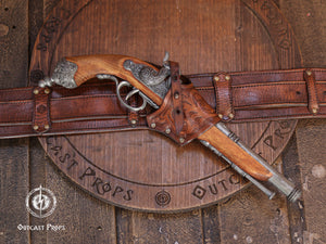 Aged brown leather flintlock holster mounted on a belt holding a pirate style flintlock pistol. Handmade leather holster with embossed tooling, brass hardware and adjustable retention strap. Designed as a cross draw holster for pirate costumes, LARP equipment, cosplay accessories and renaissance fair characters. Suitable for medium sized flintlock props while maintaining authentic historical inspired aesthetics.