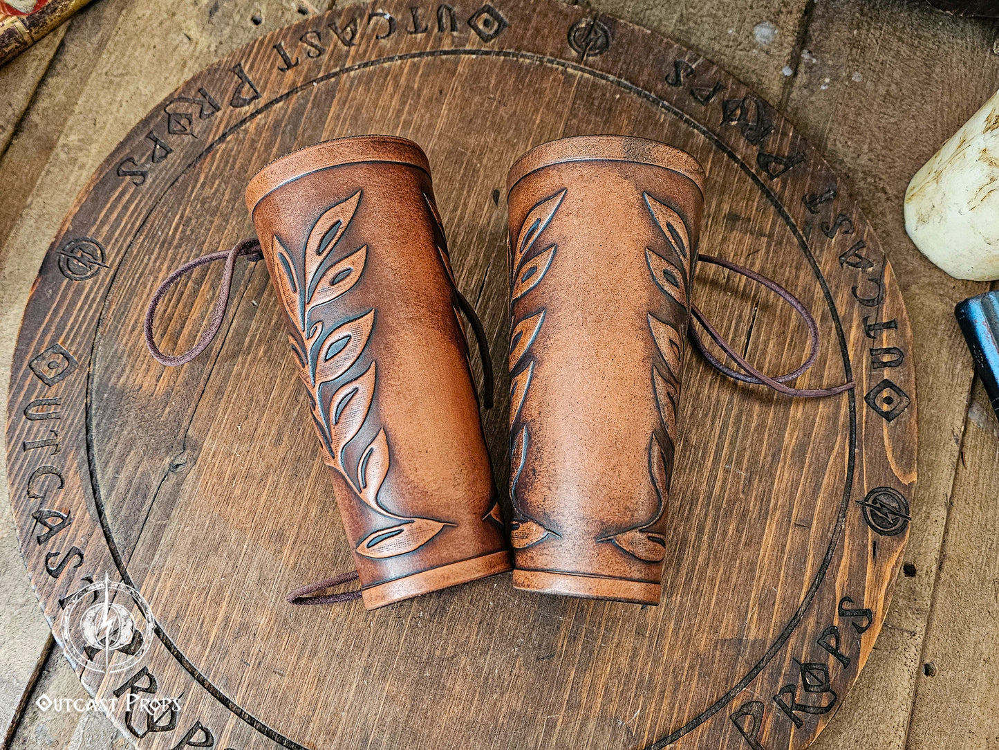Two matching brown leaf elven bracers displayed on a carved wooden board with the Outcast Props logo. Their embossed leaf pattern and aged leather finish show craftsmanship ideal for renaissance faire costumes, larp characters, elves, witches, herbalists or fae themed outfits. These leather bracers also complement roman laurel or Caesar cosplay thanks to the leaf motif. Laced fastening and warm tones highlight their elegant look.