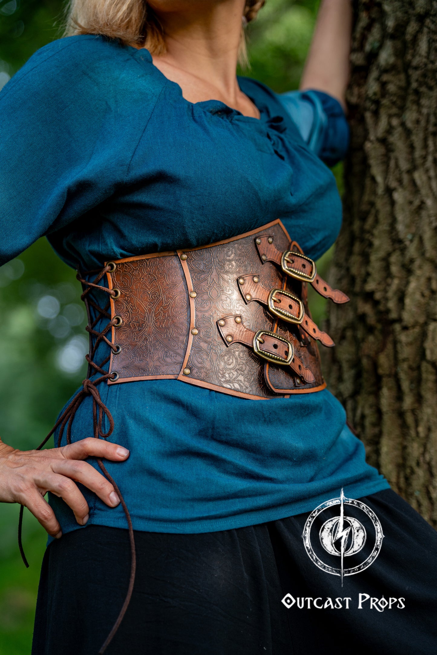 Person wearing a blue top and brown leather corset with decorative elements, standing against a tree. Embroidered Leather Underbust Corset – Elegant Brown Waist Cincher for Noble LARP Outfits, Steampunk and Fantasy Glamour