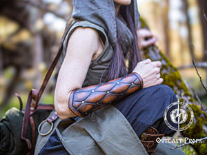 A person wearing a quilted patterned leather bracer with metal buckles and straps, seated in a wooden forest setting. Falken leather bracers for noble costume LARP outfit medieval fantasy accessories for Ren Faire cosplay arm guards for knight character gear
