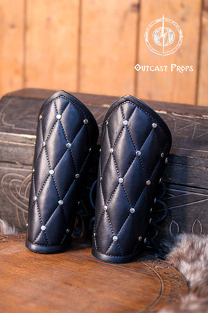 A person wearing a quilted patterned leather bracer with metal buckles and straps, seated in a wooden forest setting. Falken leather bracers for noble costume LARP outfit medieval fantasy accessories for Ren Faire cosplay arm guards for knight character gear