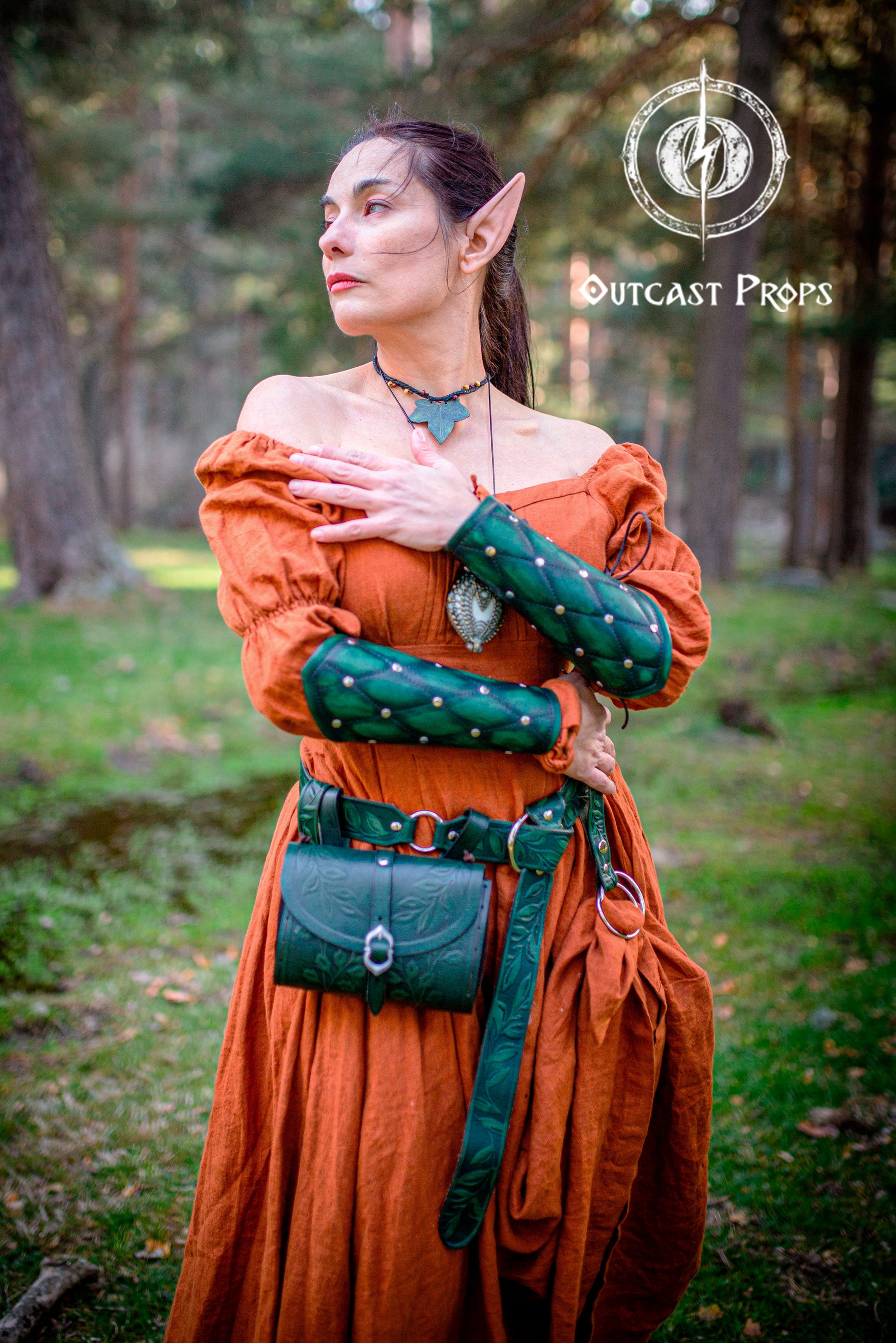 A fantasy elf cosplayer stands proudly in a forest, wearing a Renaissance fair outfit with a green Floral Leather Belt with O-Rings, matching hip bag, and green leather bracers. Her orange dress and pointy ears evoke a vivid fantasy world. This look is perfect for Fantasy Elven Waist Belt, Ren Faire Medieval Cosplay Belt, and LARP Costume Gift handmade belt enthusiasts looking for epic festival outfits.