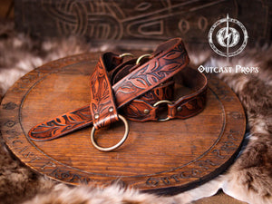 A handcrafted Floral Leather Belt with O-Rings in rich brown tones, curled elegantly atop a rustic carved wooden board. This Renaissance Leather Belt with O Rings Fantasy Elven Waist Belt is tooled by hand with intricate leaf designs, ideal for medieval cosplay, LARP outfits, and fantasy fairs. Made with premium vegetable-tanned leather, showcasing artisanal craftsmanship and durable beauty.