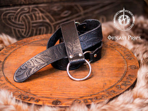Black Floral Leather Belt with O-Rings coiled neatly on a carved medieval-style wooden platform, surrounded by faux fur props. Ideal for Renaissance Leather Belt with O Rings, Fantasy Elven Waist Belt, and LARP Costume Gift handmade belt fans. Featuring a classic hand-tooled leaf design and sturdy vegetable-tanned leather, this belt blends dark fantasy aesthetics with authentic craftsmanship.