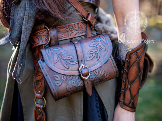 A woman wearing a brown floral hip bag stands in a natural forest setting. The bag hangs from a matching floral belt, paired with a shoulder strap and a leather arm cuff. The embossed leather shines in the sunlight, ideal for ren faire, viking cosplay, or adventurer outfits. Brown floral leather hip bag for LARP or cosplay, perfect for rogue characters, traveling merchants or fantasy warriors. Elven Leather Hip Bag Gift for Renaissance Faire Floral Belt Pouch Medieval LARP