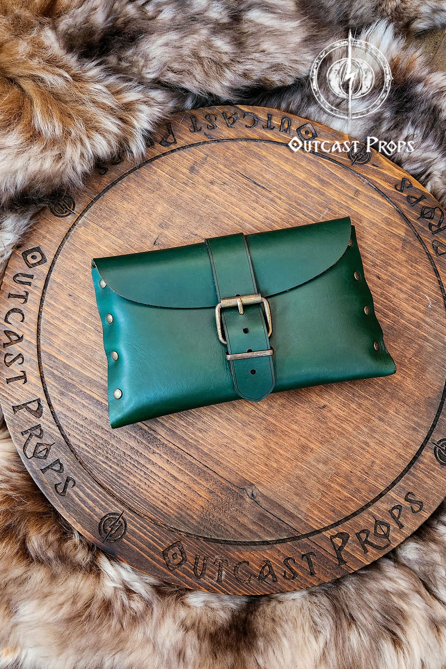 Green leather LARP pouch with brass buckle displayed on wood board marked Outcast Props. Surrounded by brown fur, this handmade medieval belt pouch features visible rivets and soft shine from polished leather. Suitable for Renaissance fair, Celtic, or fantasy costumes. Clear alt description for blind users and SEO long tails: green leather fantasy pouch.