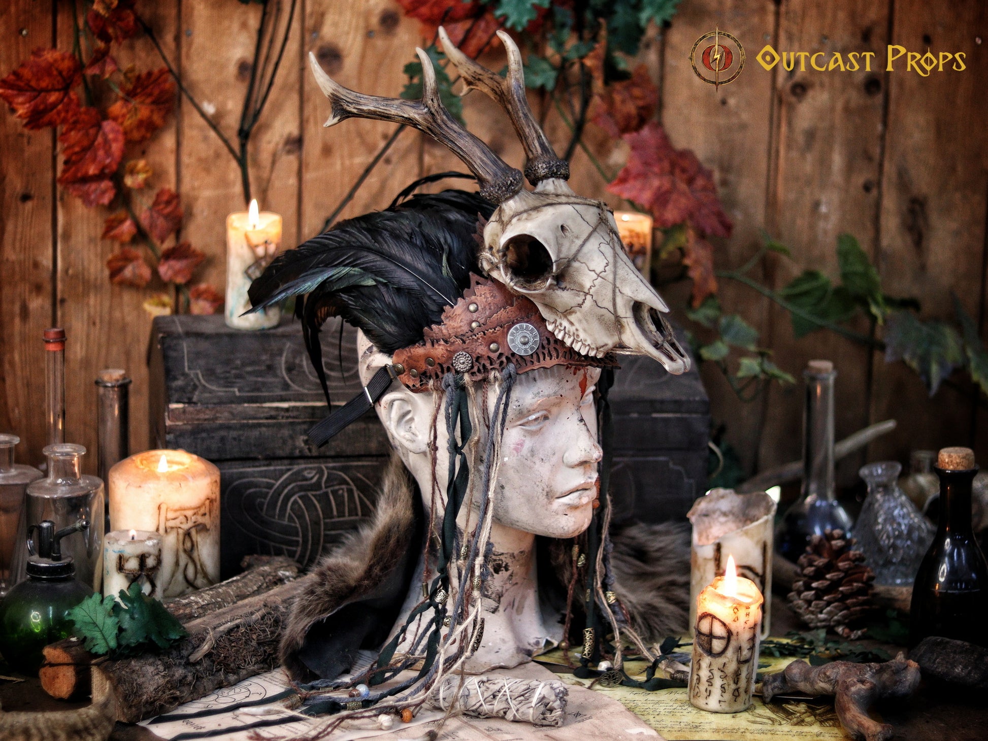 A Celtic adorned tiara with a resin skull, deer horns, feathers, bone pieces, beads, and fangs displayed on a table with a wooden background and candles. Beltane Headpiece tribal deer horns skull and feathers tiara leather and resin