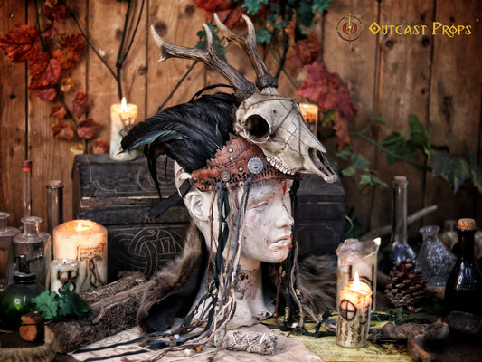 A Celtic adorned tiara with a resin skull, deer horns, feathers, bone pieces, beads, and fangs displayed on a table with a wooden background and candles. Beltane Headpiece tribal deer horns skull and feathers tiara leather and resin
