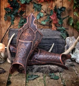 Artisanal Celtic greaves displayed on a wooden stage surrounded by Viking props, antlers, and rune candles. The brown leather leg armor features Nordic knotwork and a dragon design. Handmade by Outcast Props for fantasy warriors, LARP adventurers, or medieval cosplay outfits.