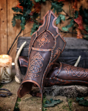 Close-up of a pair of leather greaves with intricate Celtic knotwork and Norse dragon design, aged with Outcast Props' epic painting techniques. These leg armors are ideal for Viking LARP characters, Celtic cosplay, or fantasy costumes. Made from vegetable-tanned leather with hand-applied rivets.